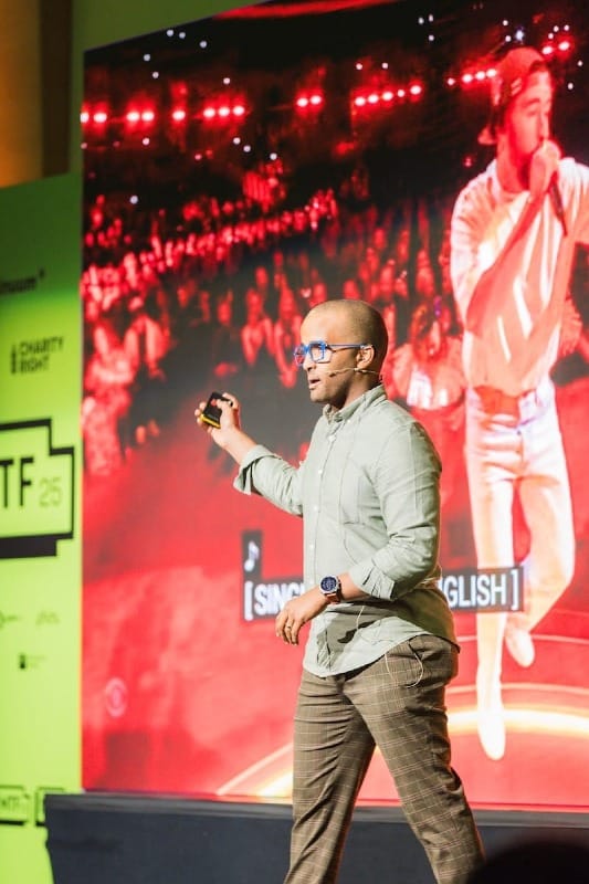 Ahmed on stage and standing in front of a screen while a clicker and speaking to an audience.